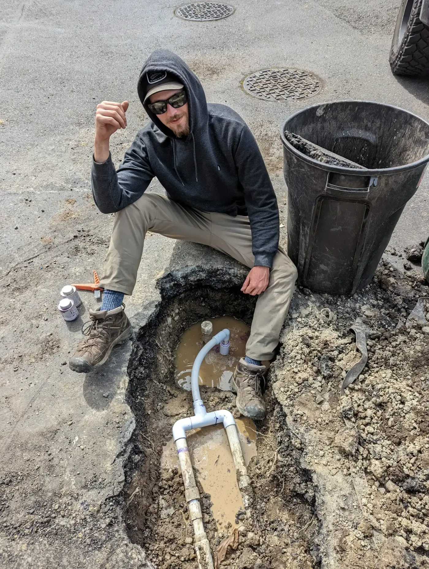 Worker beside excavated asphalt trench holding new PVC drain piping