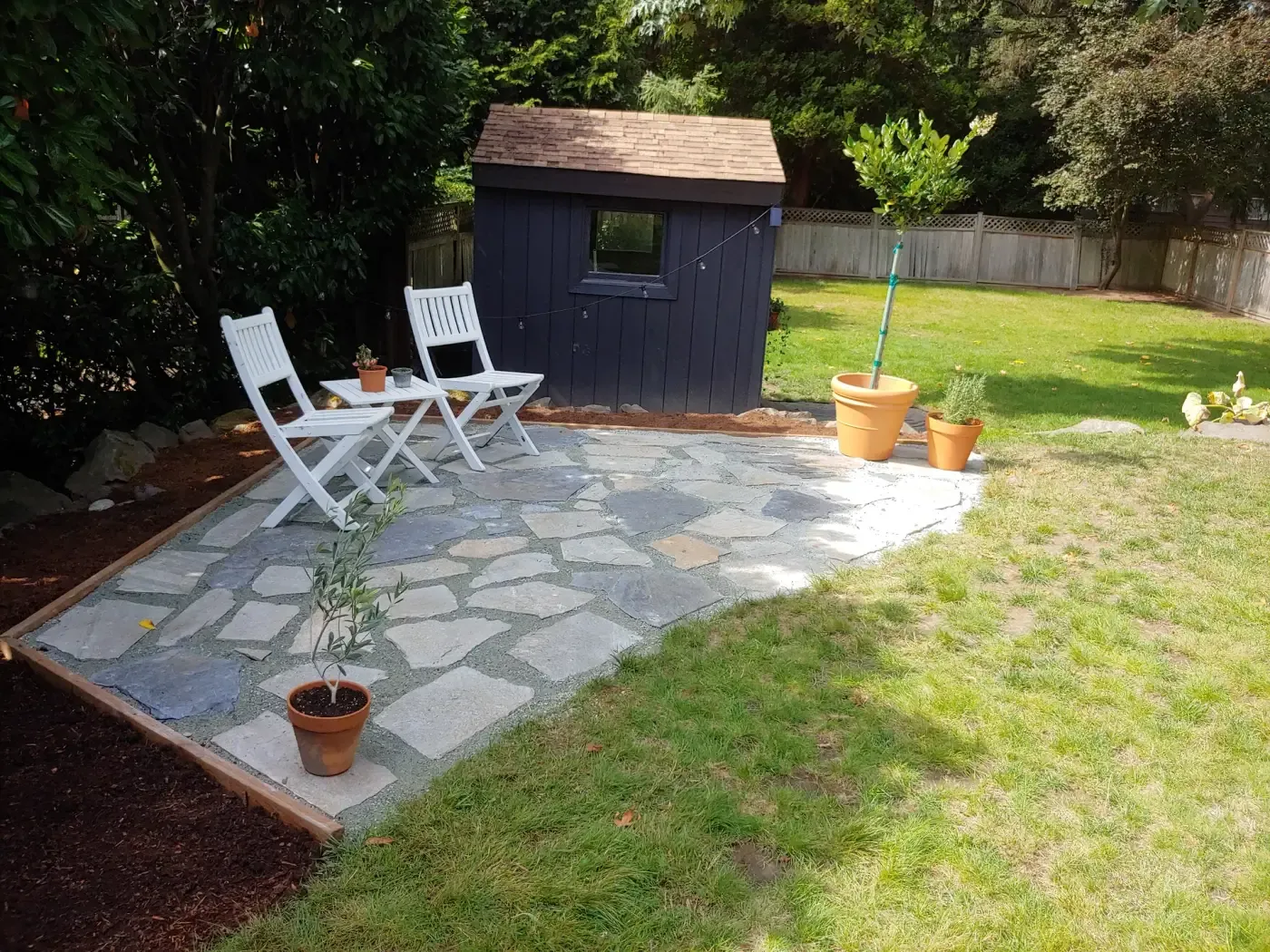 Grey flagstone patio with white chairs next to dark shed