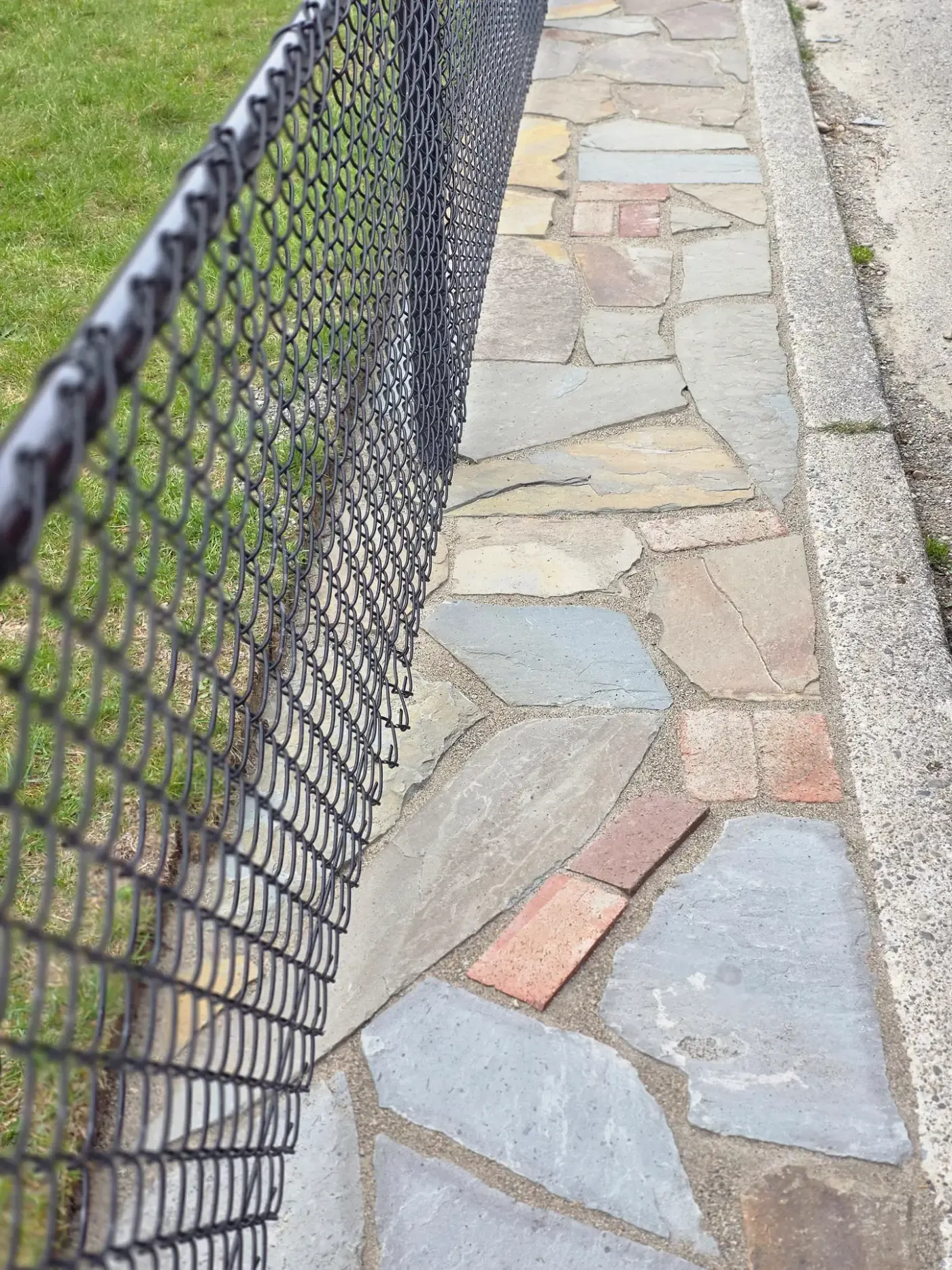 Mortared flagstone walkway with brick inlays running between a chain link fence and the street curb