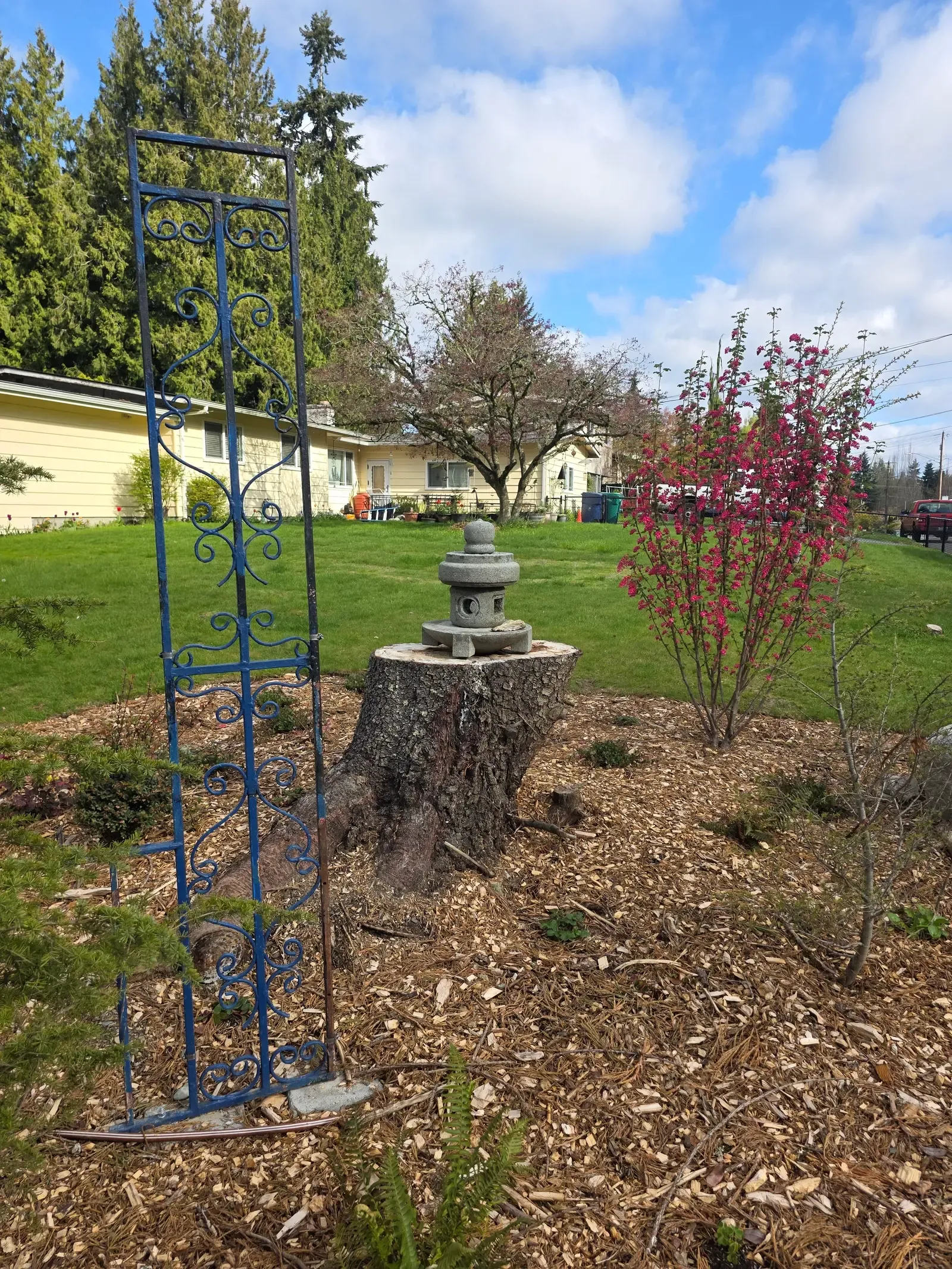 Mulched bed with new shrubs, metal obelisk and the lantern on the stump