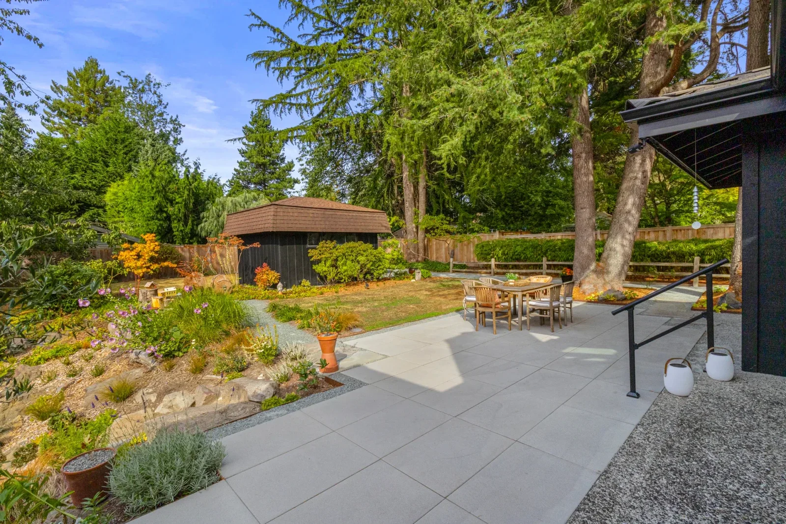 Large format slab patio with a dining set, a planted berm of perennials and a garden shed under tall firs