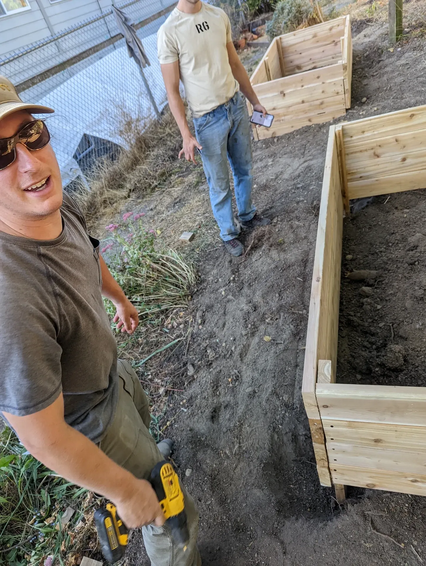 Trevor with a cordless drill beside newly built cedar planter boxes, a second crew member working behind him