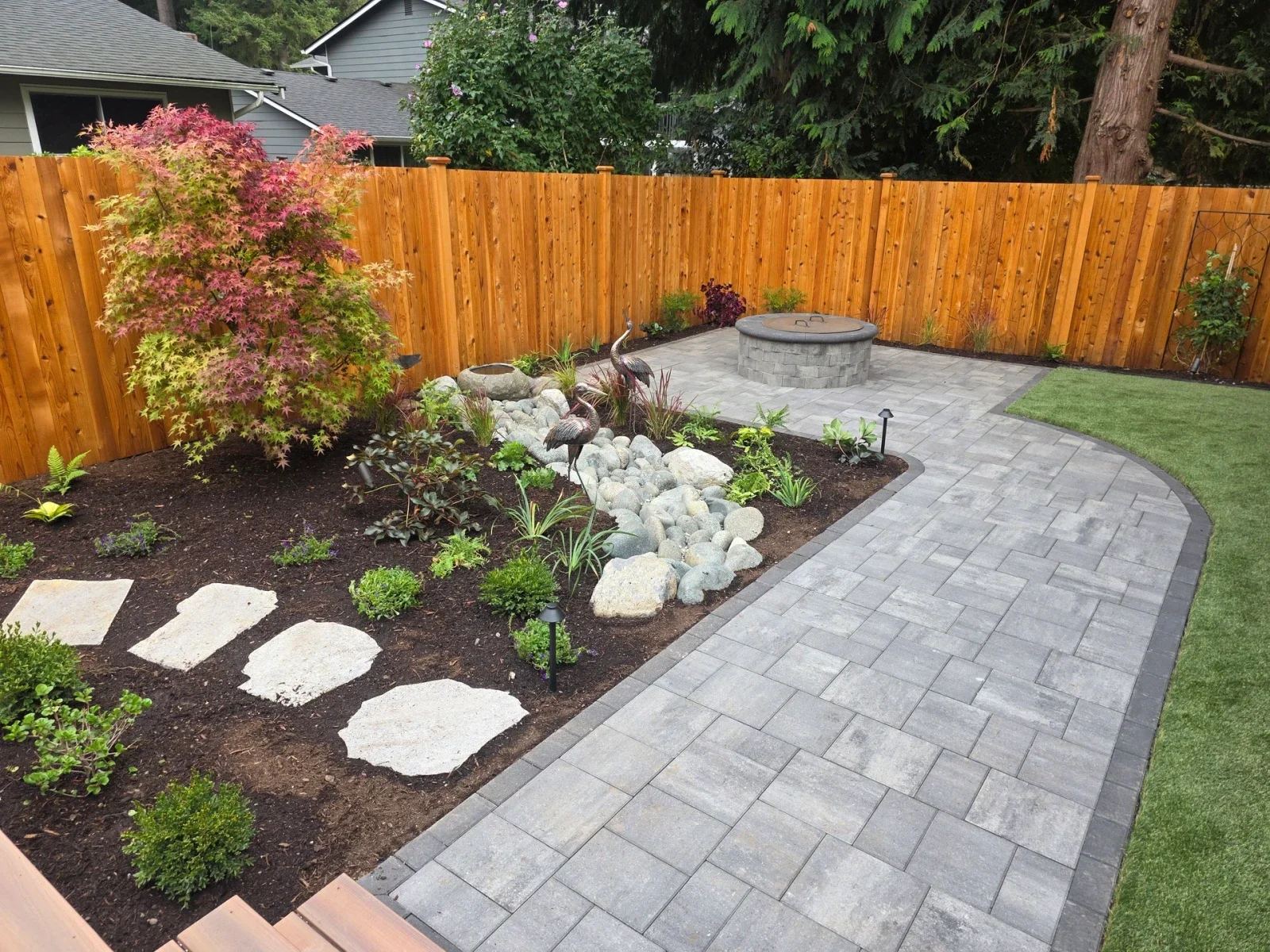Finished backyard build: grey paver patio with a dark border and a round fire pit, a boulder stream bed and stepping stones in new planting, and a new cedar fence