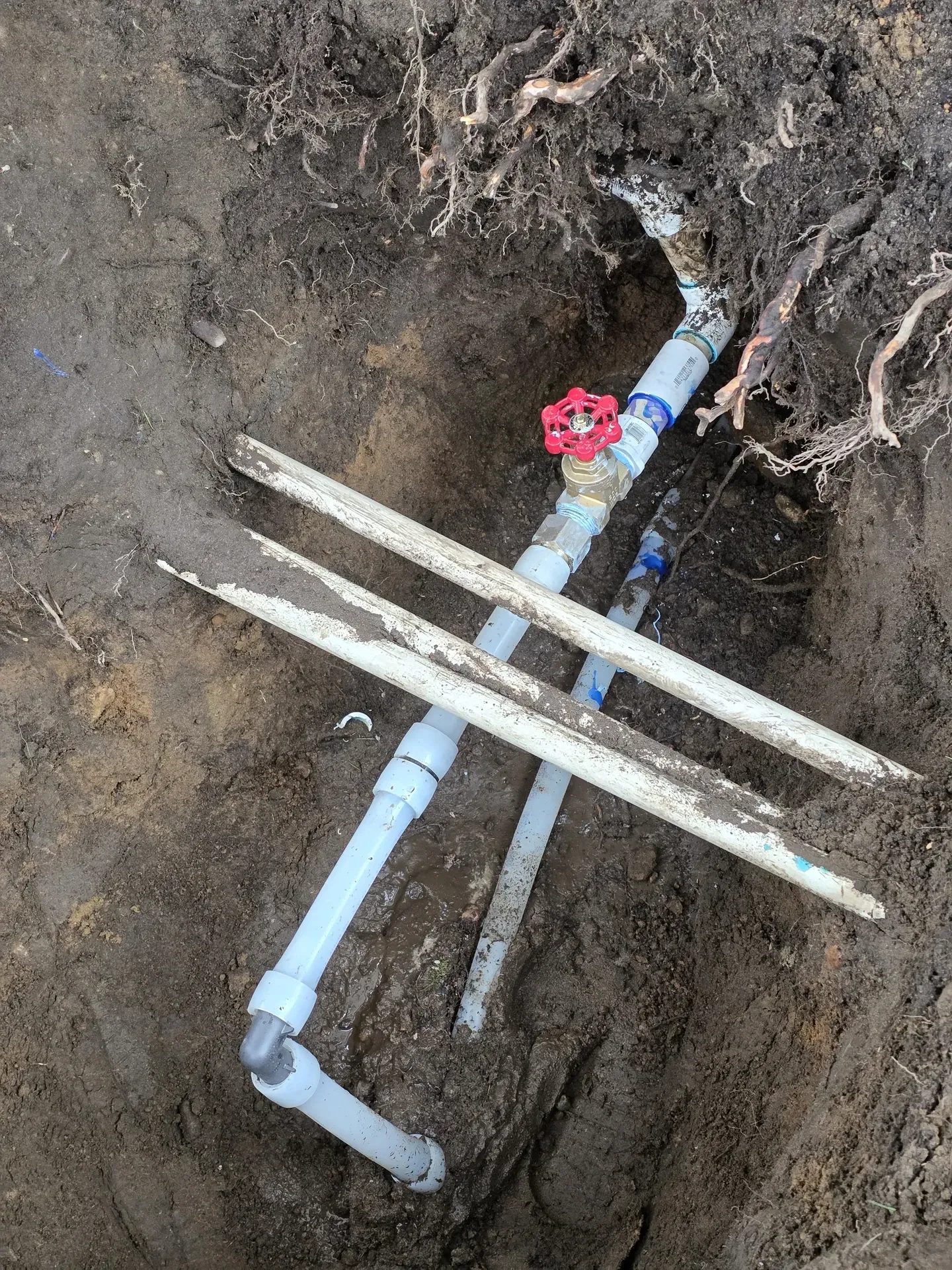 Open trench showing a PVC manifold and a red-handled shutoff valve