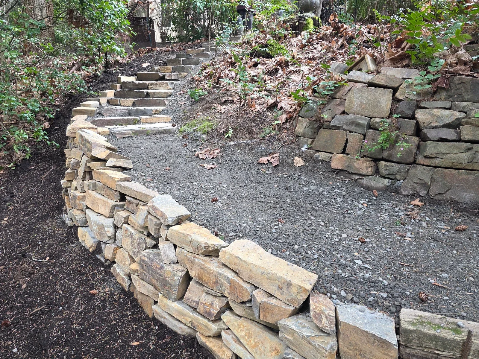 Dry stacked stone retaining wall curving below a run of stone steps on a wooded slope