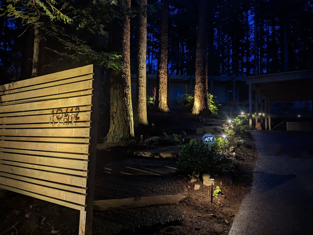 Wooden sign illuminated at night with trees and a house entrance in the background.