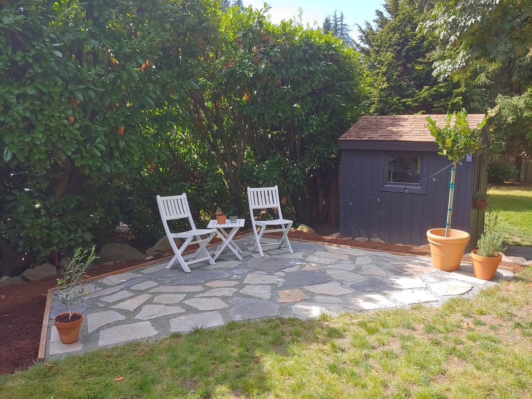 Cozy patio with two white chairs, stone flooring, potted plants, and a garden shed.