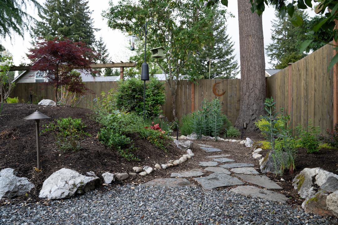 Garden pathway with stone path, lush greenery, and wooden fence in a serene outdoor setting.