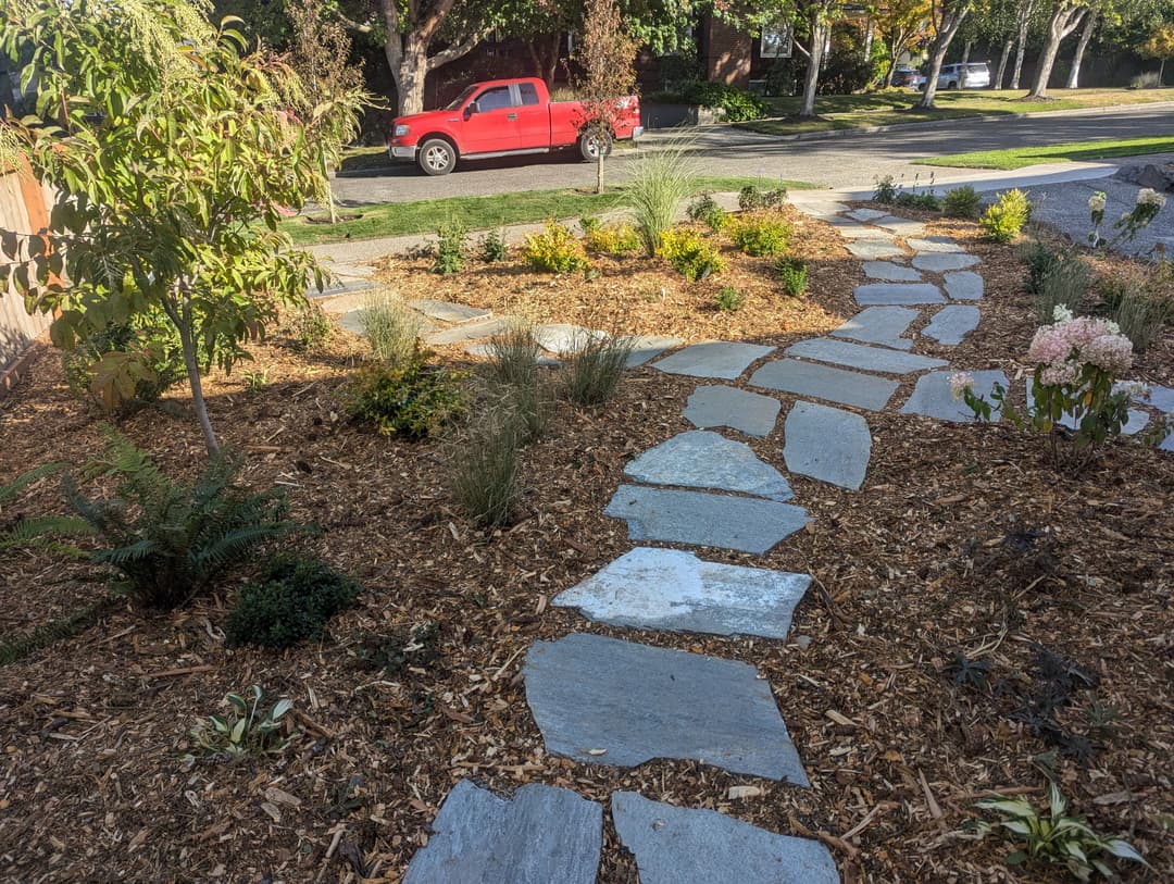 Pathway made of stone in a landscaped garden with shrubs and a red truck in the background.