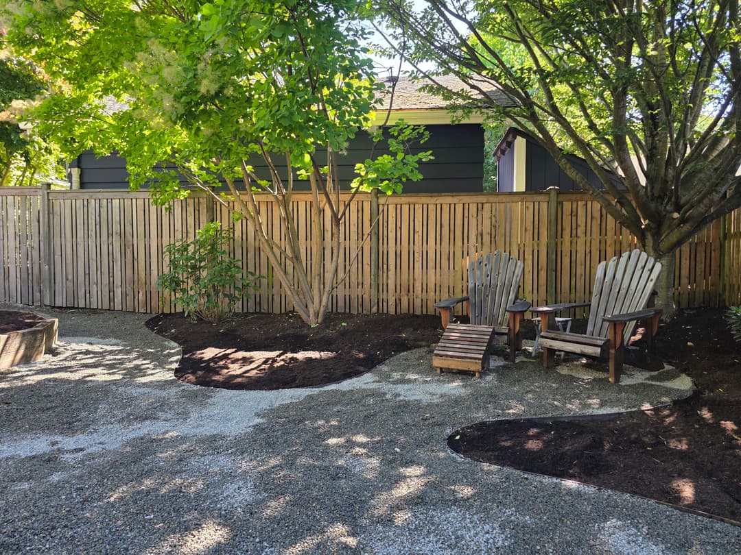 Cozy backyard retreat with gravel pathway, wooden fence, and adirondack chairs under trees.