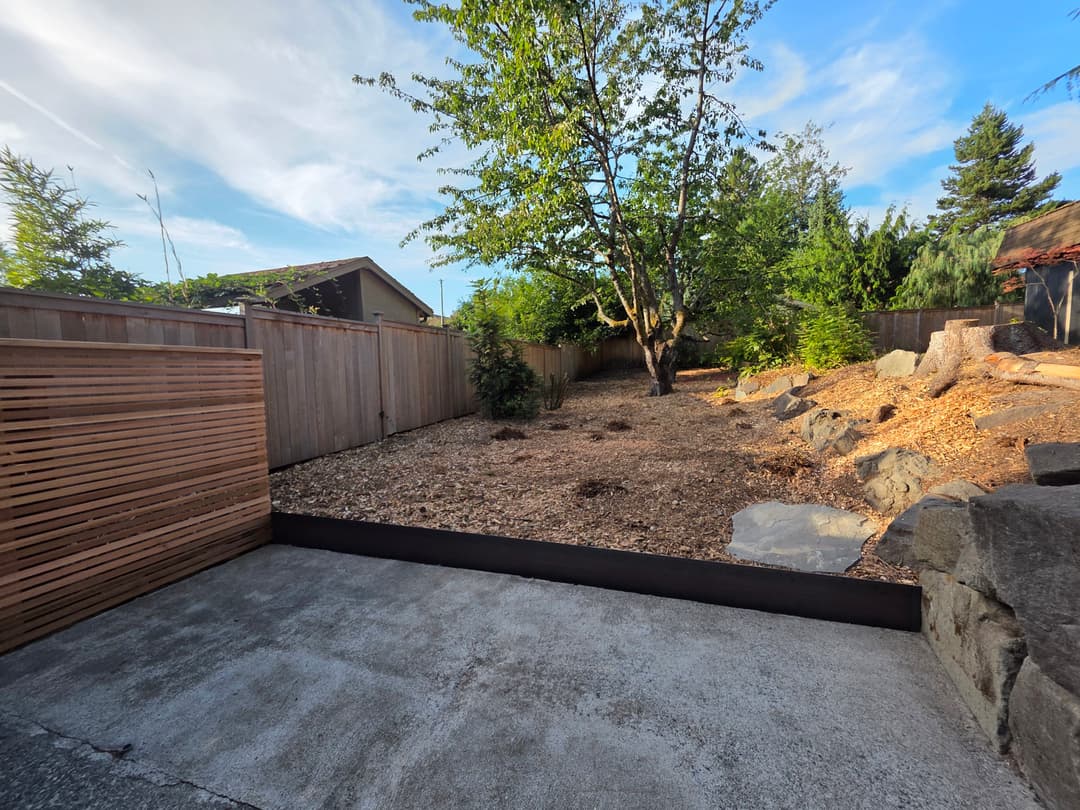 landscaped yard with a tree, wooden fence, and rocky area under a clear blue sky