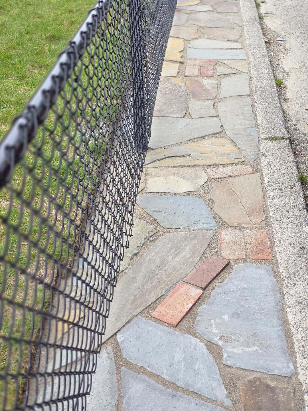 Stone pathway bordered by a black chain-link fence, with green grass nearby.