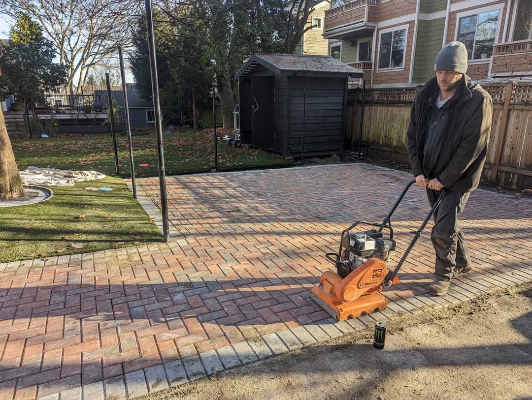 Person operating a plate compactor on a newly paved brick patio in a backyard setting.
