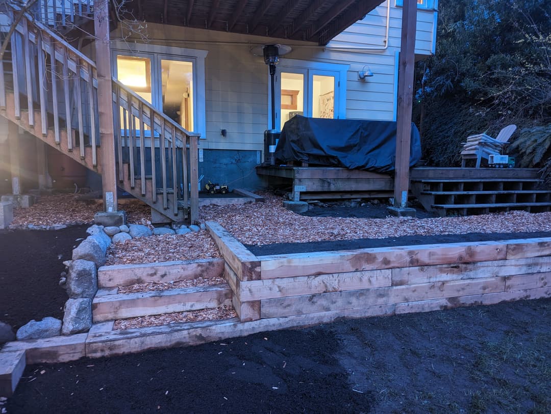 Wooden retaining wall and landscaped area with stairs leading to a deck and grill.