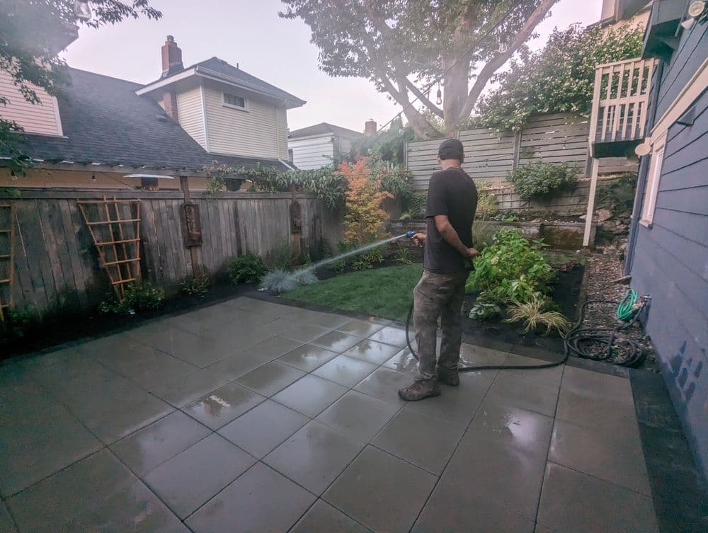 Man watering a landscaped backyard with patio and plants after rain.