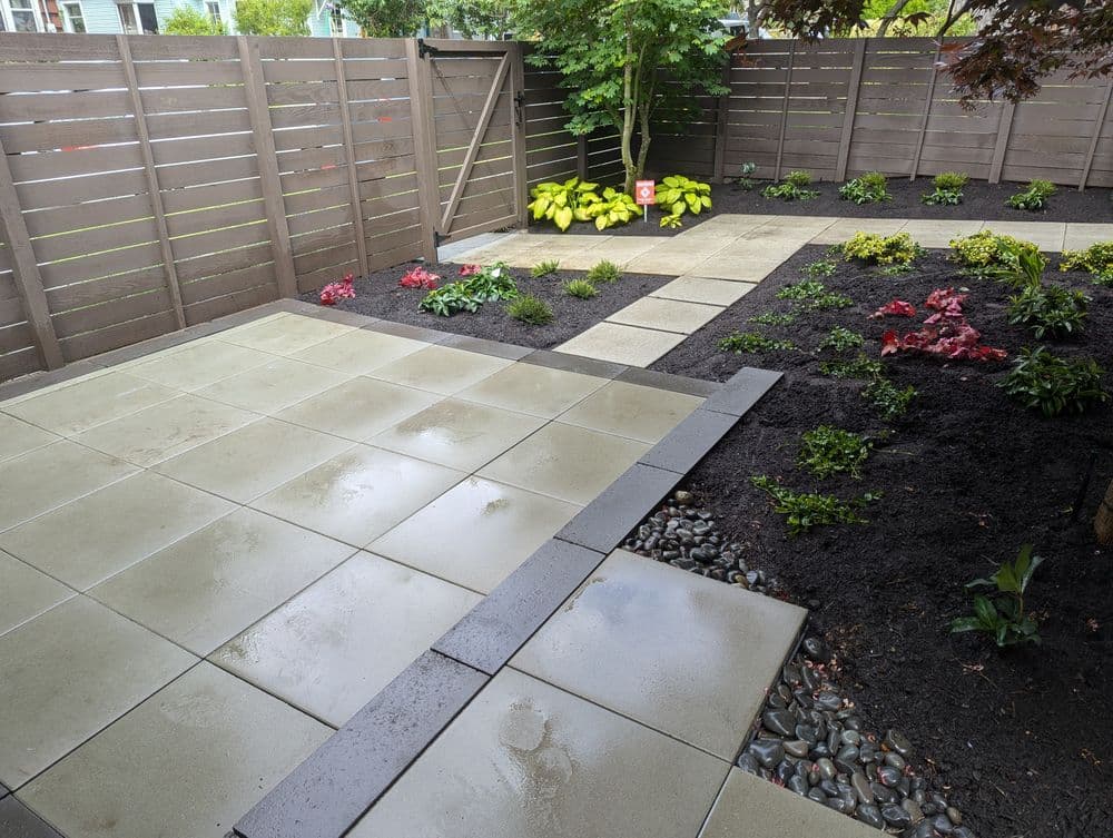 Modern backyard patio design with patterned stone tiles, greenery, and colorful flower beds.